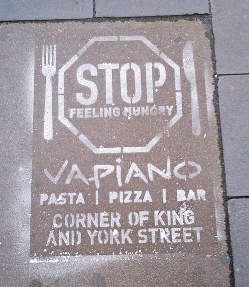 immagine fatta con stencil e spray su un marciapiede. Scritta "STOP feeling hungry", VaPiano è dietro l'angolo